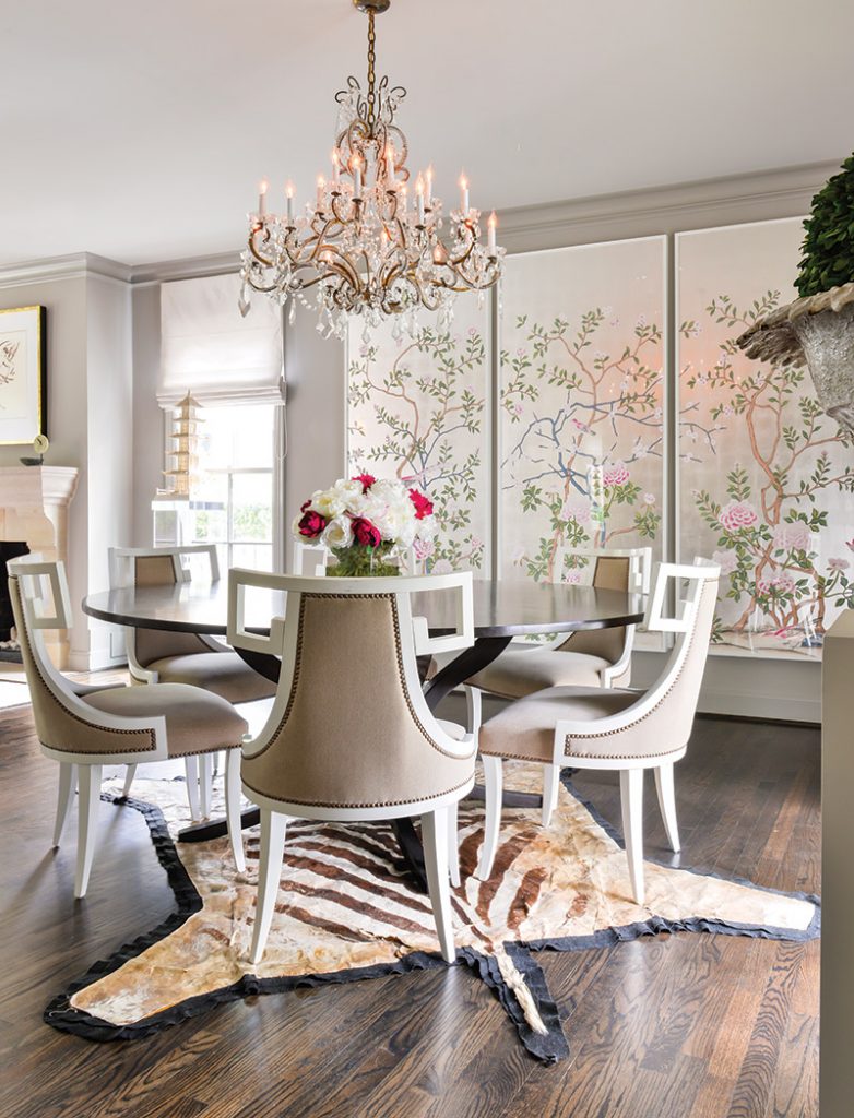 Framed panels of de Gournay on silver tea leaf paper infuse the dining room with artistic elegance, while an antique French crystal chandelier shines above a custom dining table surrounded by contemporary chairs in traditional upholstery and nail-head trim. A zebra rug adds a hint of pattern pizzazz.