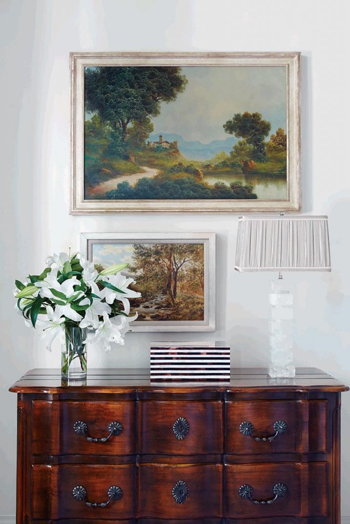 Walnut chest topped with lilies and a lamp below a gilt-framed pastoral landscape