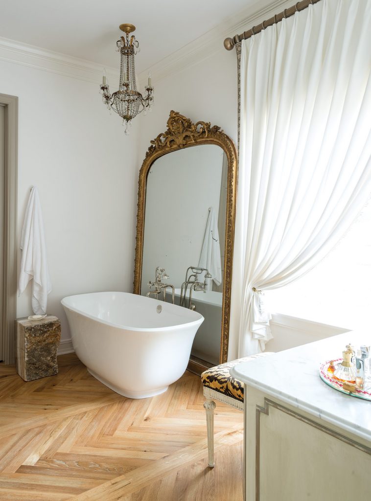 A sophisticated chevron pattern, installed by a local artist, graces the floors of the master bedroom and bath. Antiques and technology live side by side in the form of a French daybed and remote-controlled drapes. An oversized gilded mirror, billowy curtains, and a crystal-laden chandelier add to the romantic appeal of the bathroom.