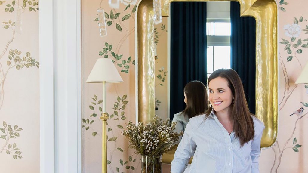 Two women in pale shirts beside a mirror in a hand-painted wallpapered room