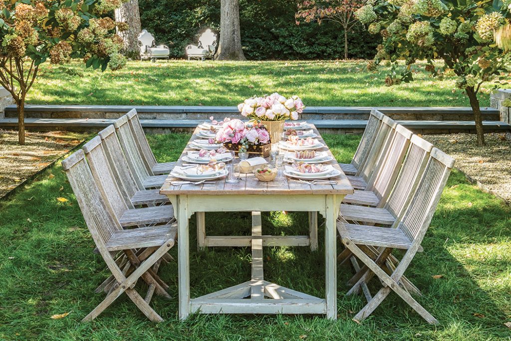 Long outdoor table set with pink flowers and folding chairs on a lawn