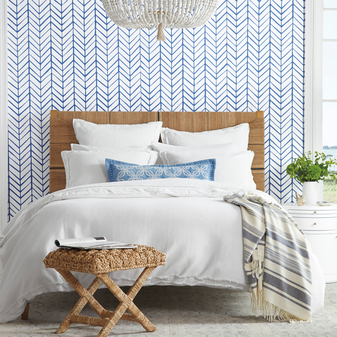 Bed with white bedding against blue chevron wallpaper and a woven bench