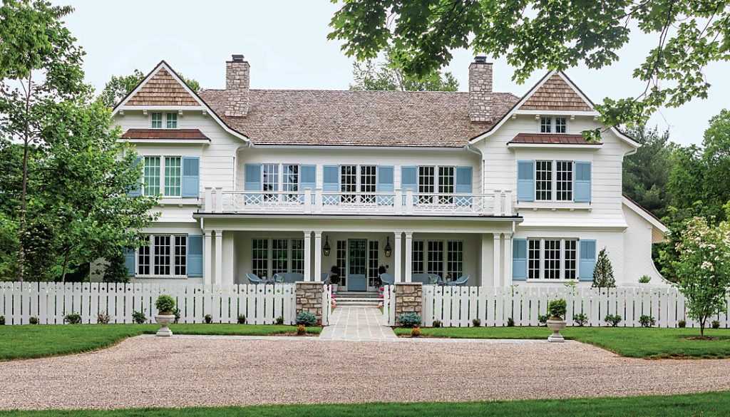 Shingled house with blue shutters behind a picket fence and gravel drive