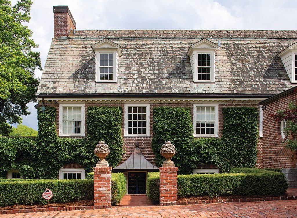 Ivy-covered brick house with dormers behind clipped hedges
