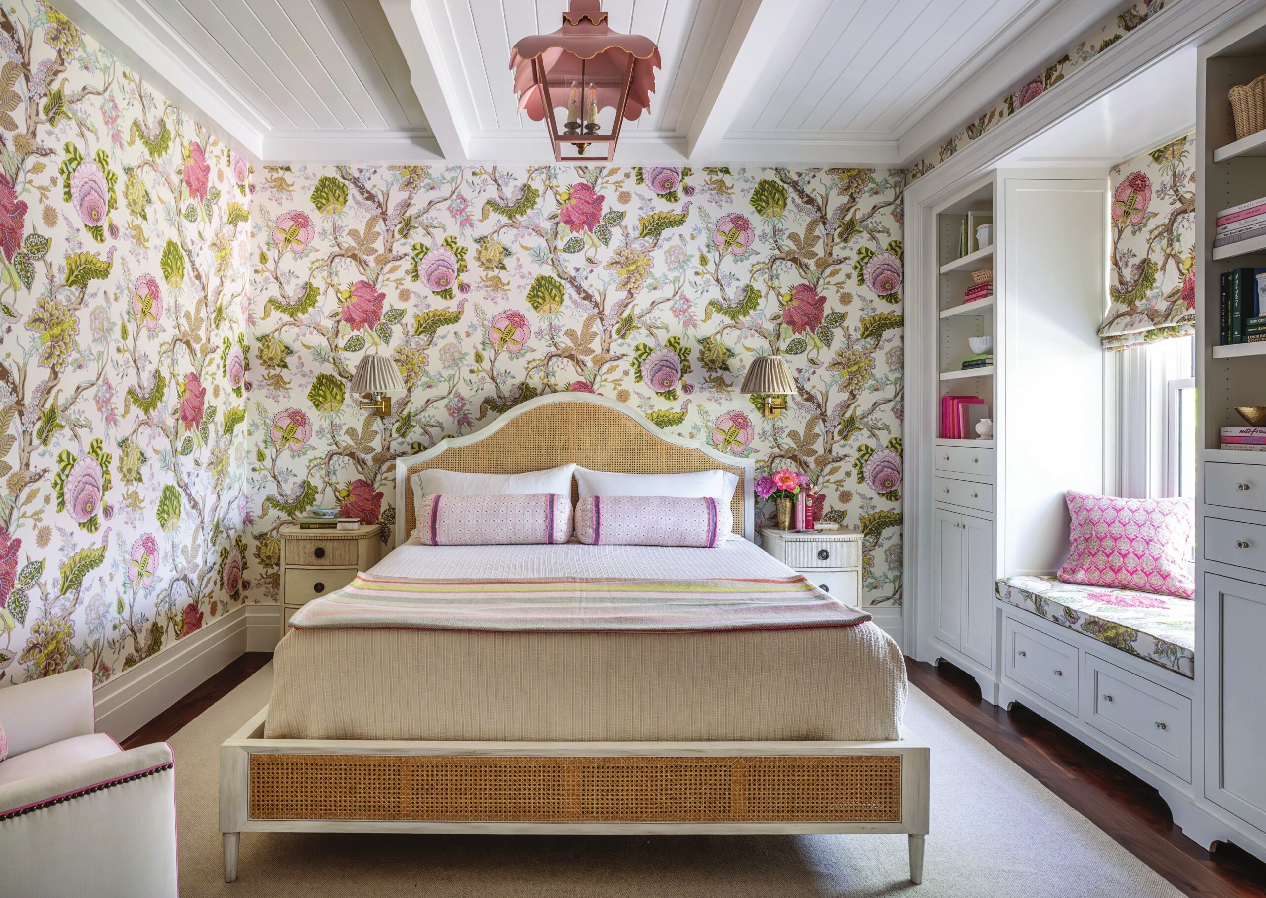 A bedroom papered in a large-scale floral, with a cane bed, pink bedding and a built-in window seat