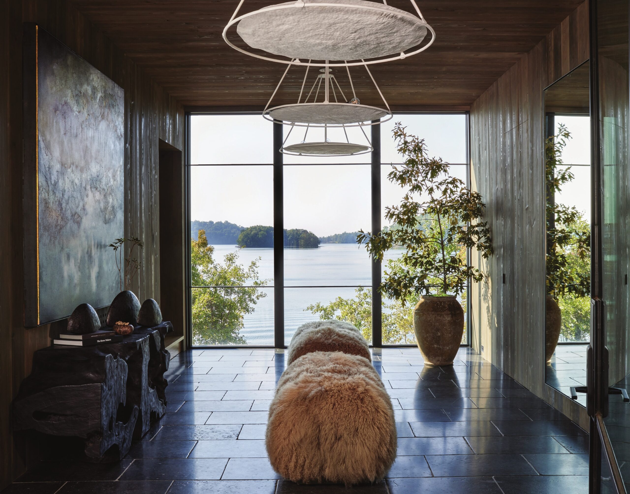 A timber and stone living room opening onto a lake, with a low bench and a view of wooded shoreline