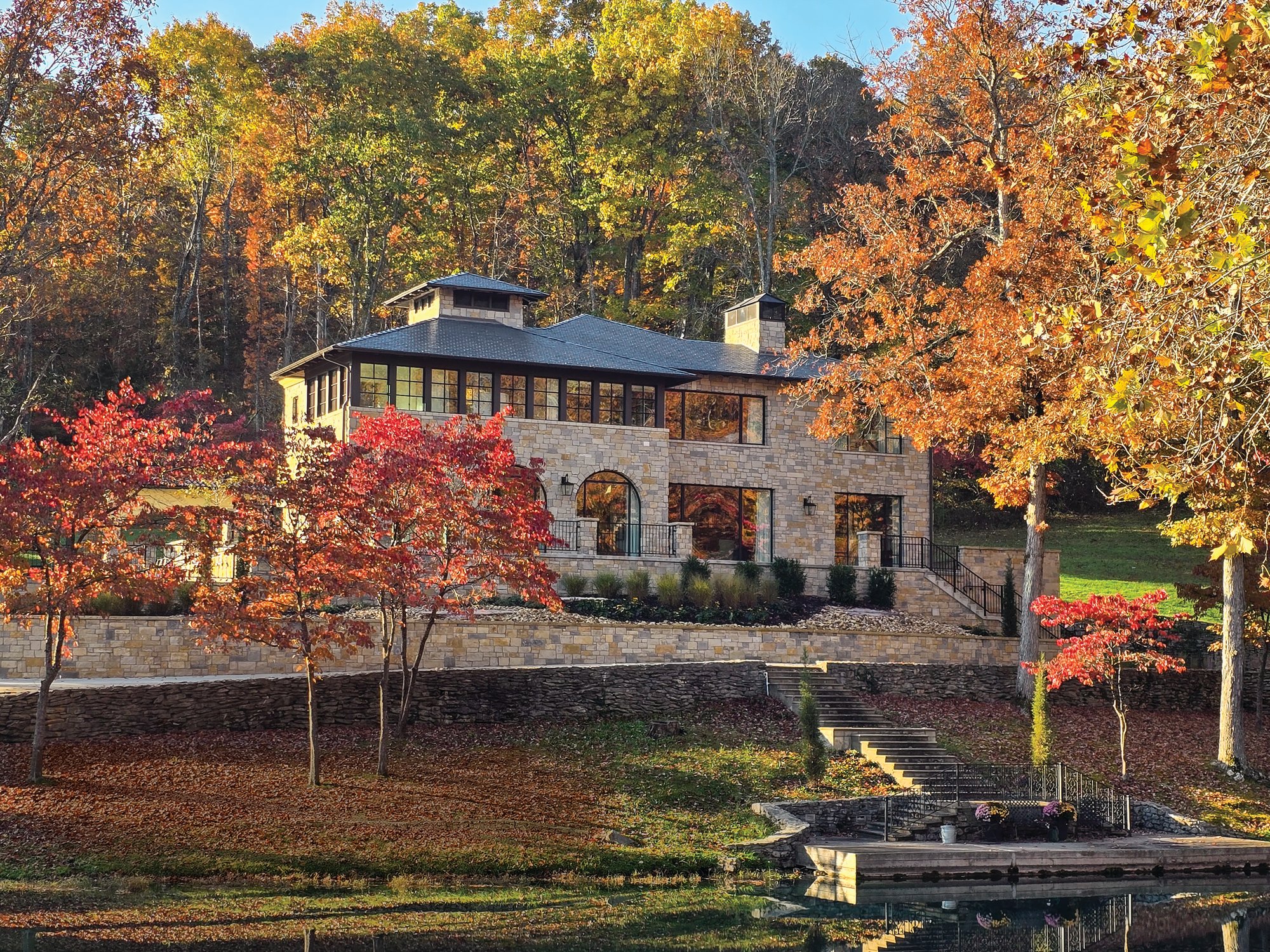 Stone facade of a new Arkansas country estate set into a wooded hillside above a spring-fed lake, framed by red and orange fall foliage