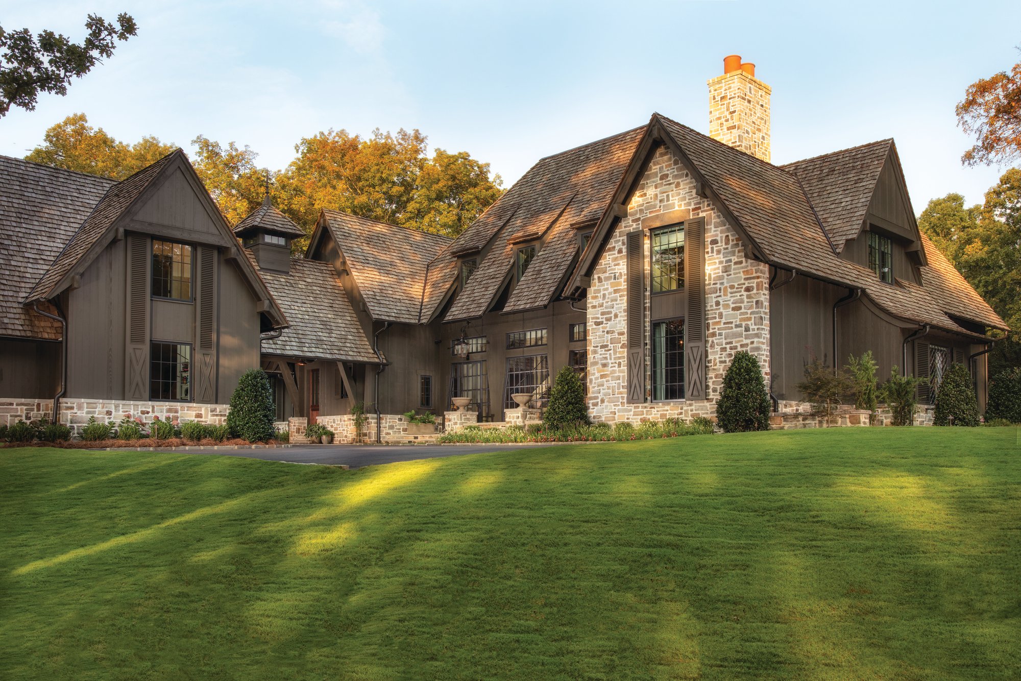 Cedar shake roof and stained wood siding on the front exterior of a timber-framed Alabama home with a stone chimney and gabled dormers at golden hour