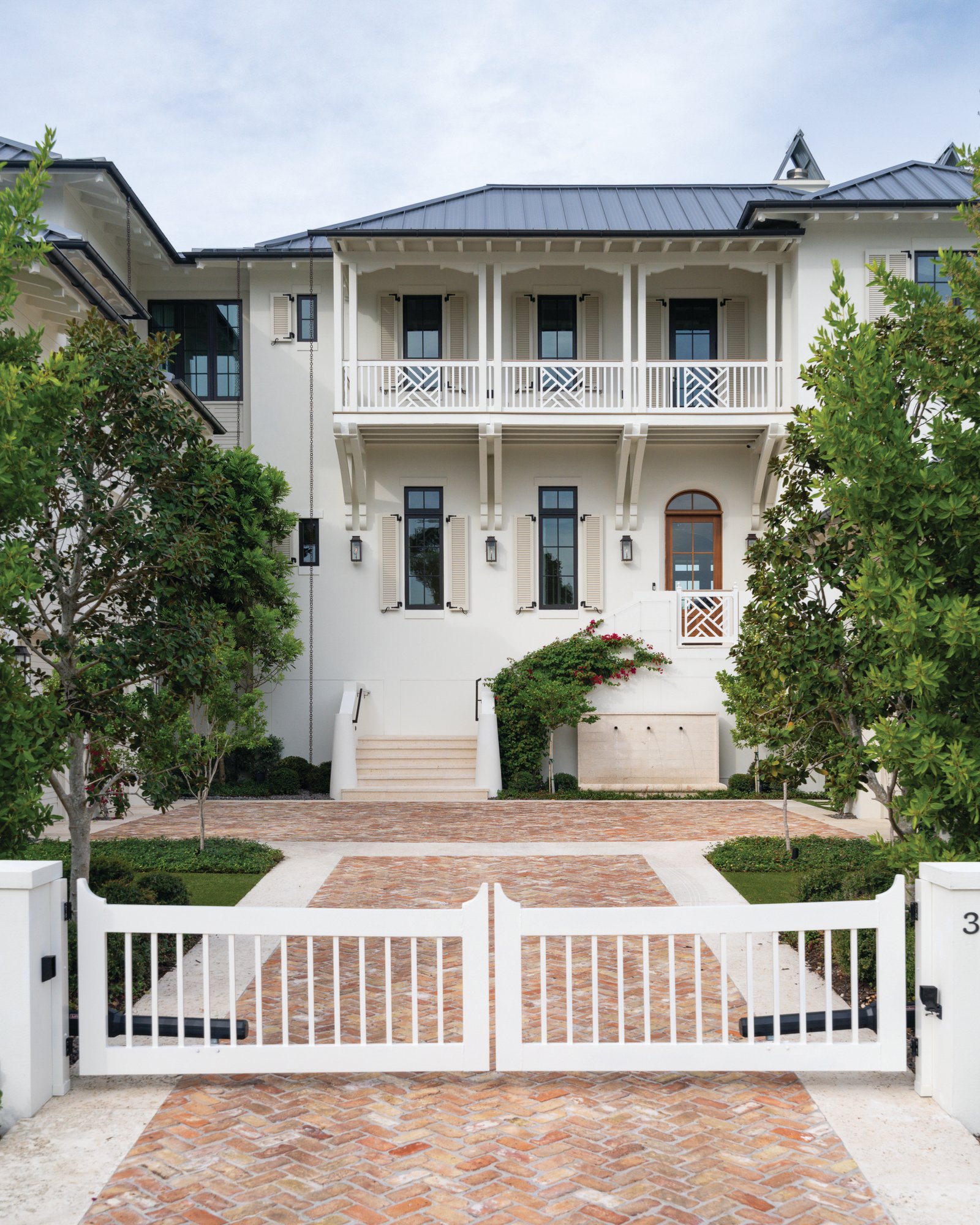 Front exterior of a white British West Indies-style home on Tampa Bay with a dark metal roof, louvered shutters, Chippendale porch railings, a herringbone brick drive, and a stone wall fountain