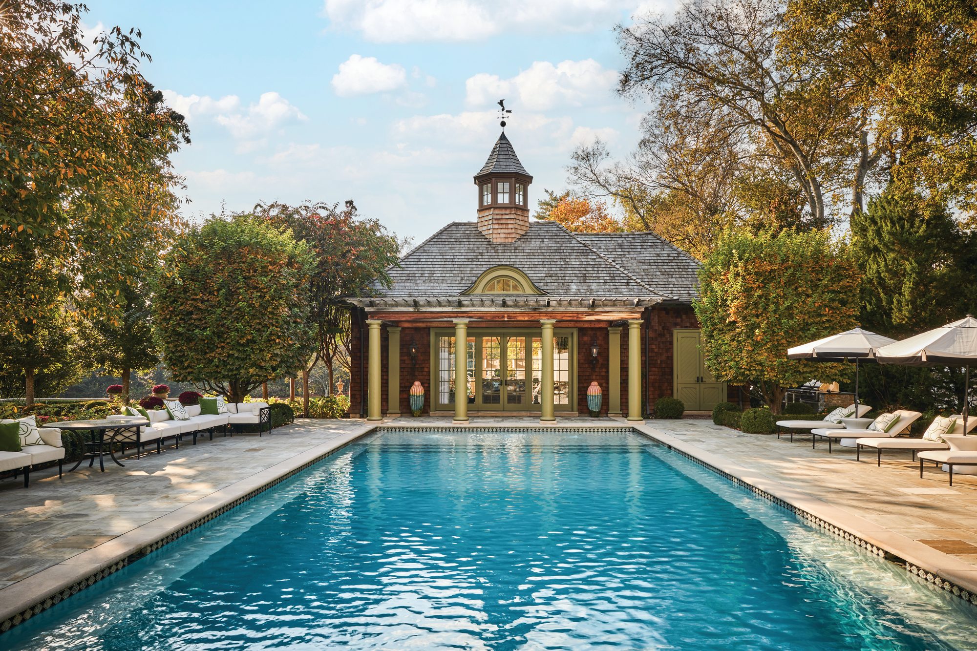 Rectangular pool with a sun deck and loungers in front of a brick pool house with green columns and a cupola