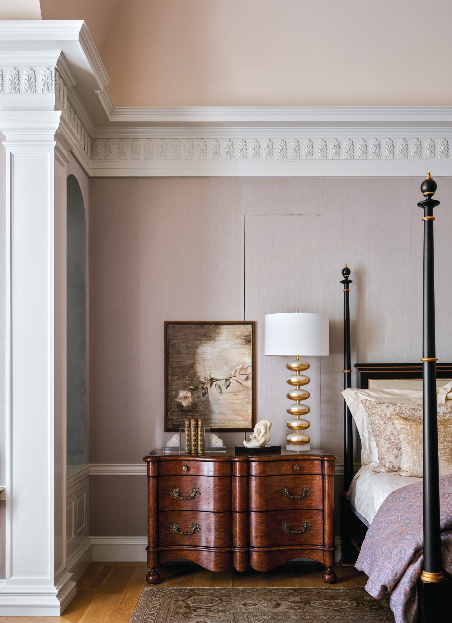 Primary bedroom with soft blush fabric-covered walls, a four-poster bed, and an antique chest topped with a lamp