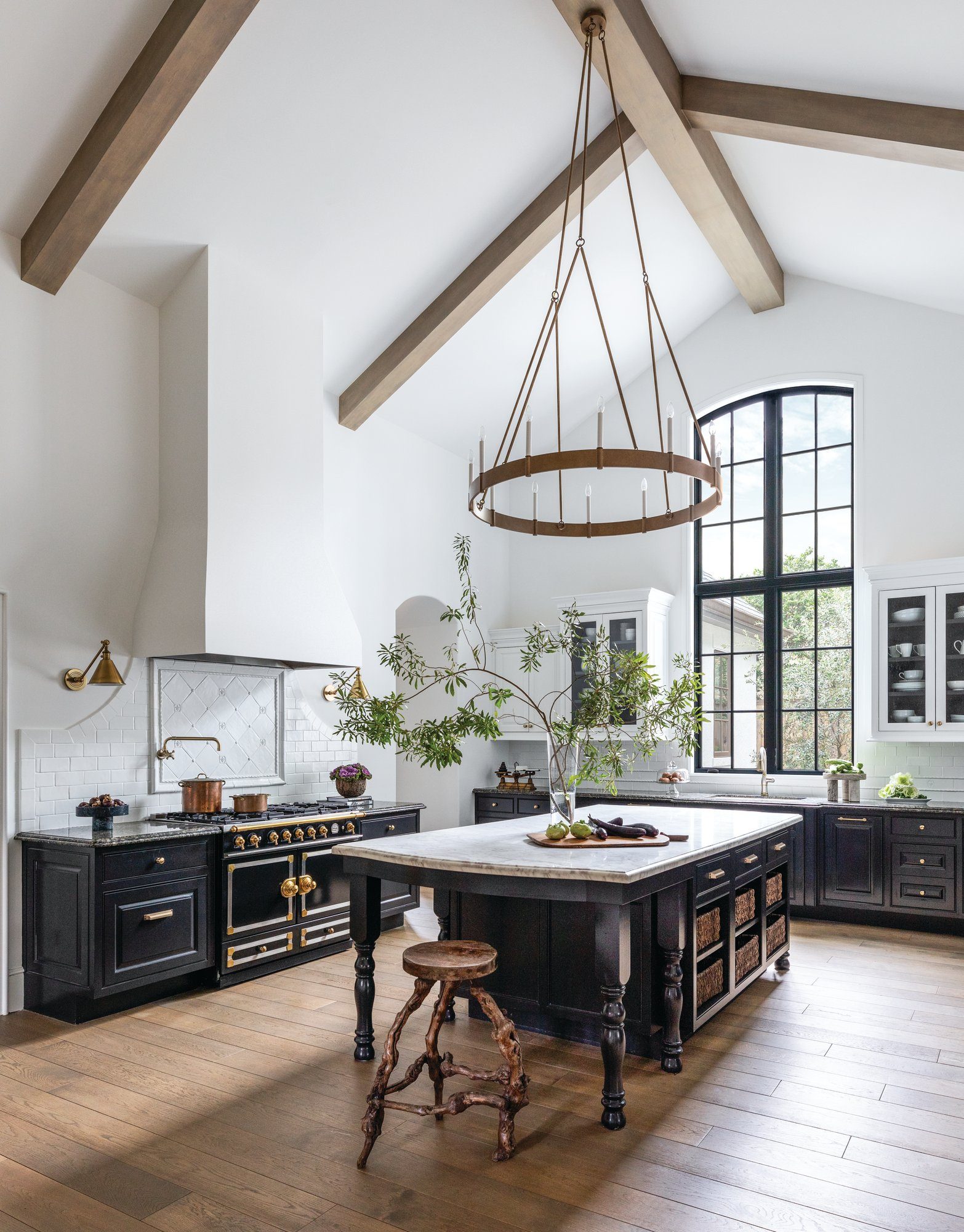 Kitchen with a vaulted beamed ceiling, black cabinetry, a marble range hood, a brass ring chandelier, and a wood island with turned legs