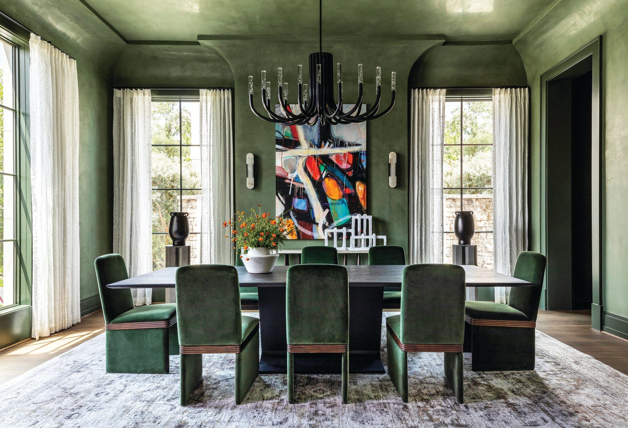 Dining room with deep green lacquered walls, a coved ceiling, green velvet chairs around an oval table, and a colorful abstract painting