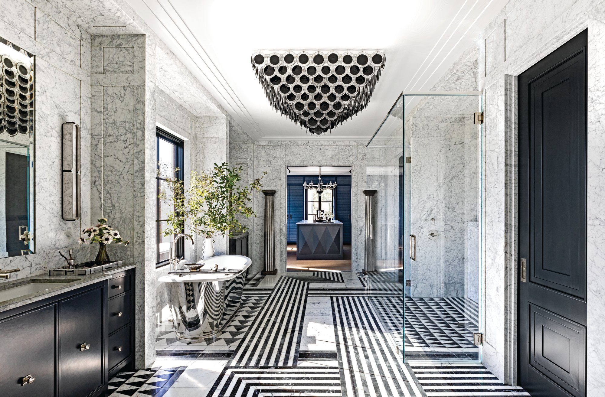Primary bath clad in marble with black-and-white striped stone floors, a black orb chandelier, a double vanity, and a freestanding tub