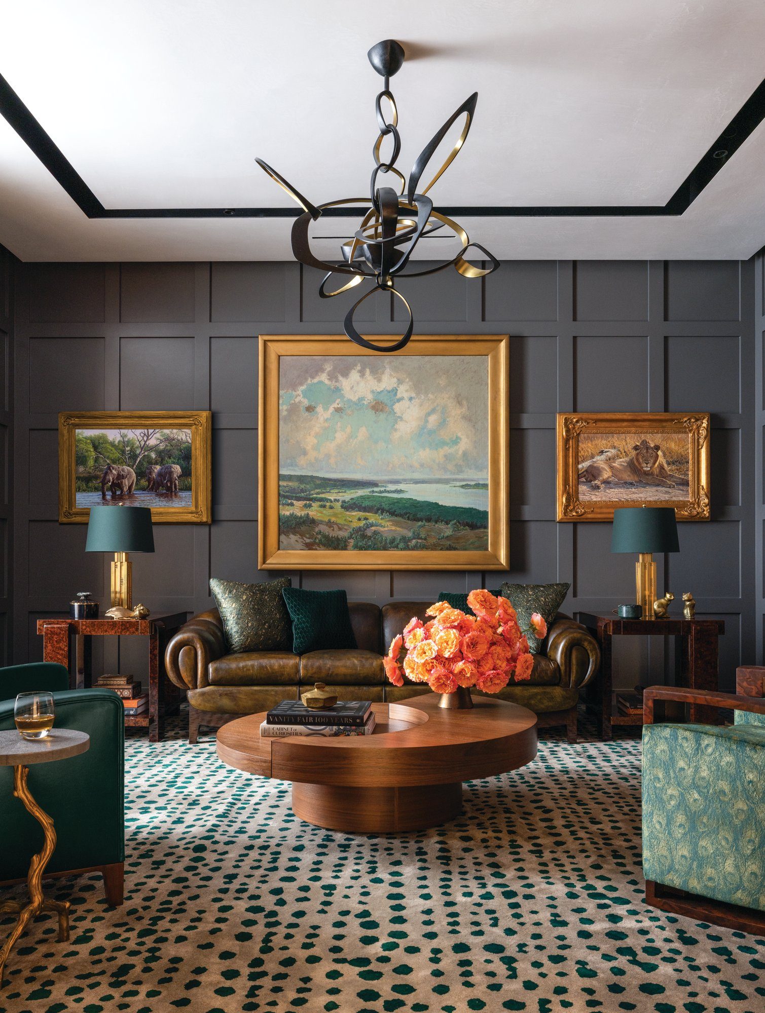 Den with charcoal board-and-batten paneling, framed landscape paintings, a green velvet sofa, a round wood table, and a spotted rug