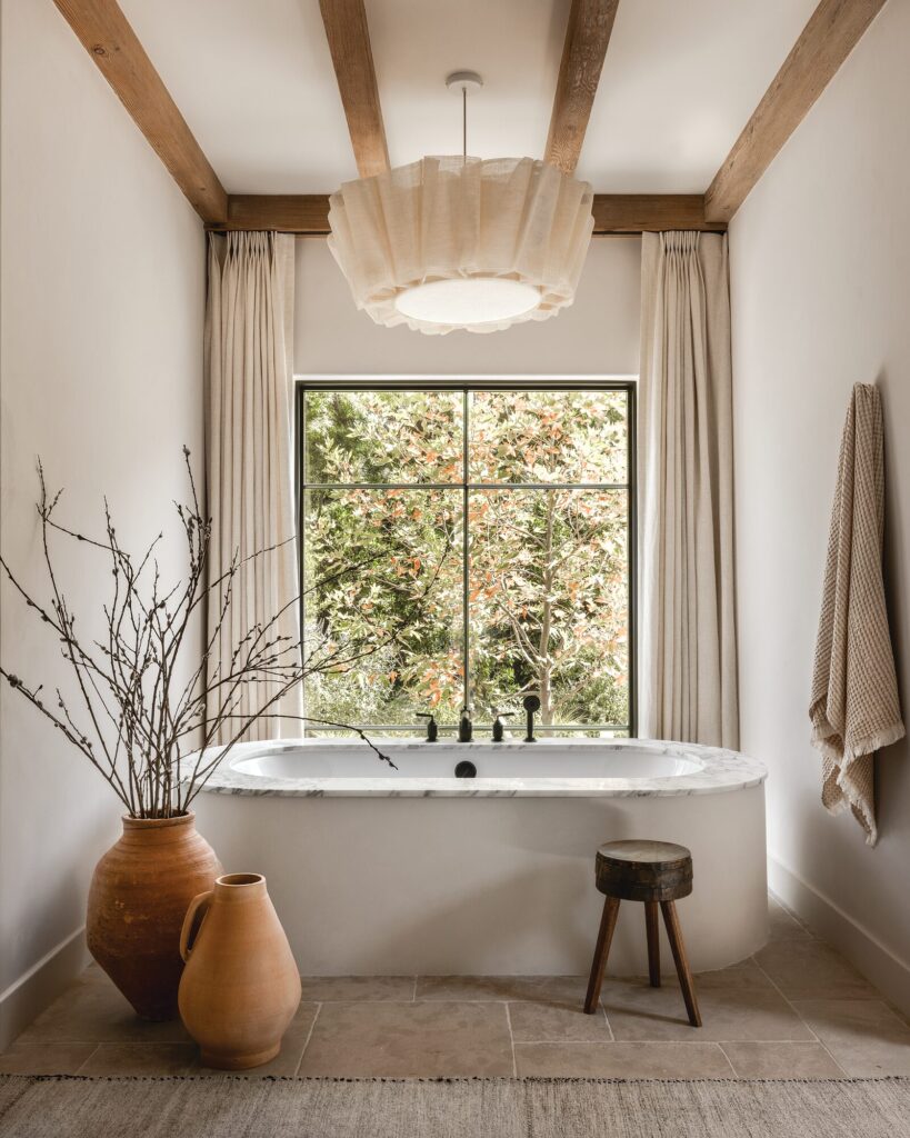 Bathroom by Amber Interiors with a freestanding tub, wood beams, terracotta vessels, and a ruffled pendant