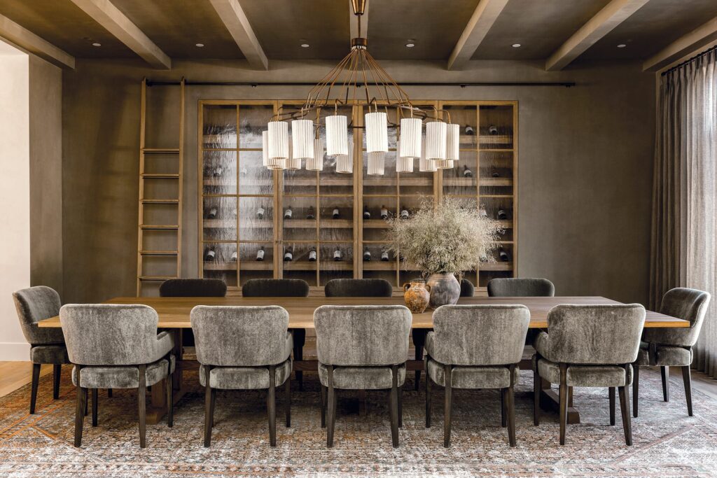 Dining room by Amber Interiors with a long wood table, upholstered chairs, and a wall of display shelving