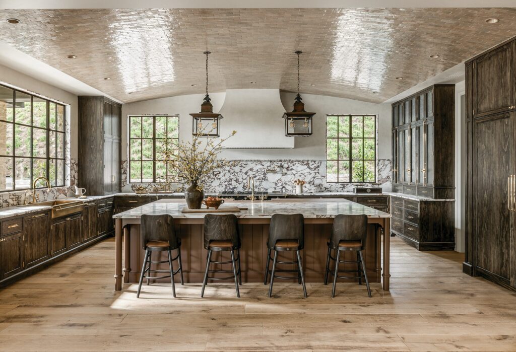 Kitchen by Amber Interiors with dark wood cabinetry, a marble backsplash, lantern pendants, and a large island