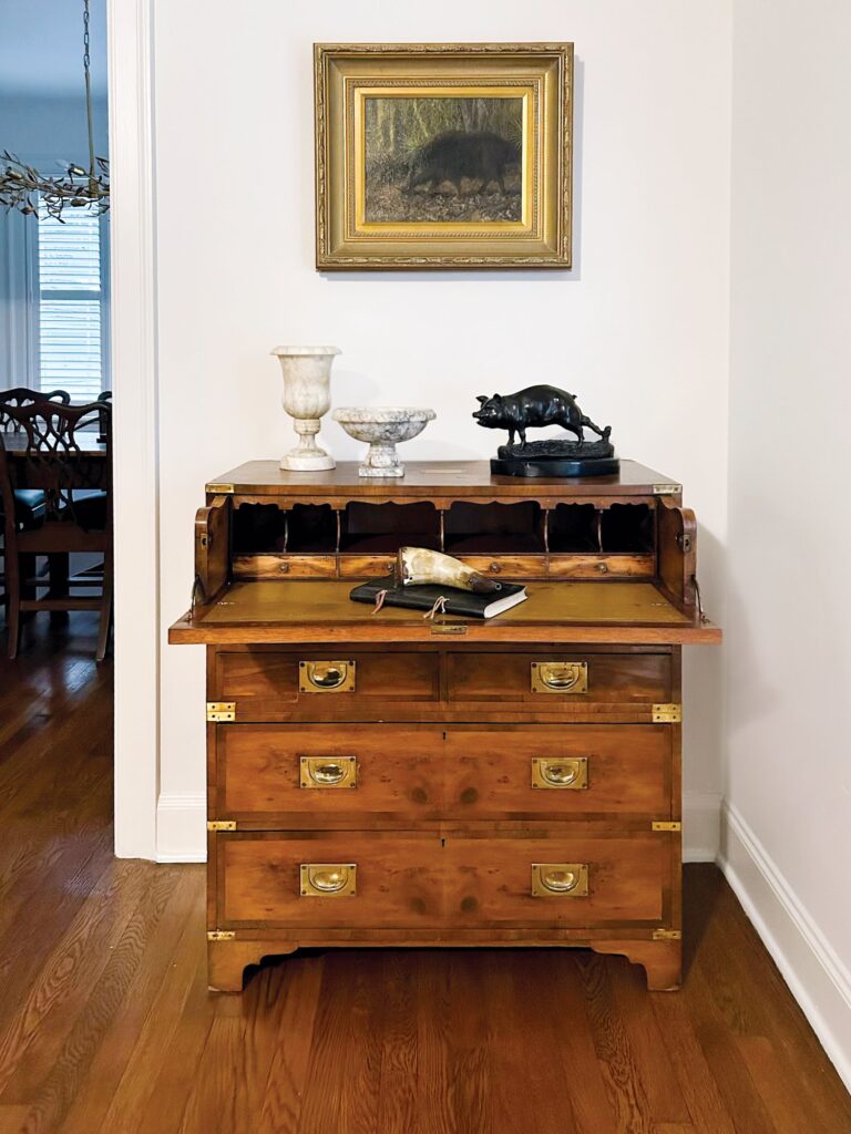 Campaign-style secretary chest with an open fall-front writing surface, interior pigeonholes, and brass recessed pulls, beneath a framed landscape painting
