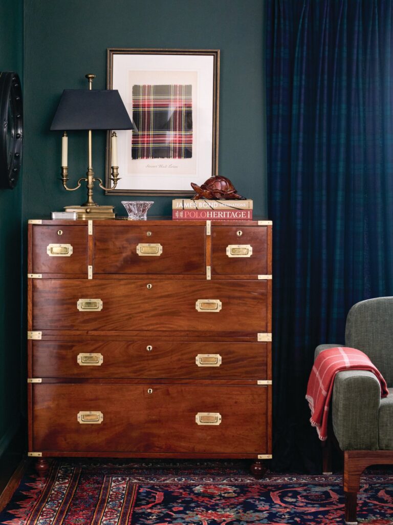 Nineteenth-century English mahogany campaign chest of drawers with brass corner guards and recessed handles, styled with a brass lamp and plaid art against a dark green wall