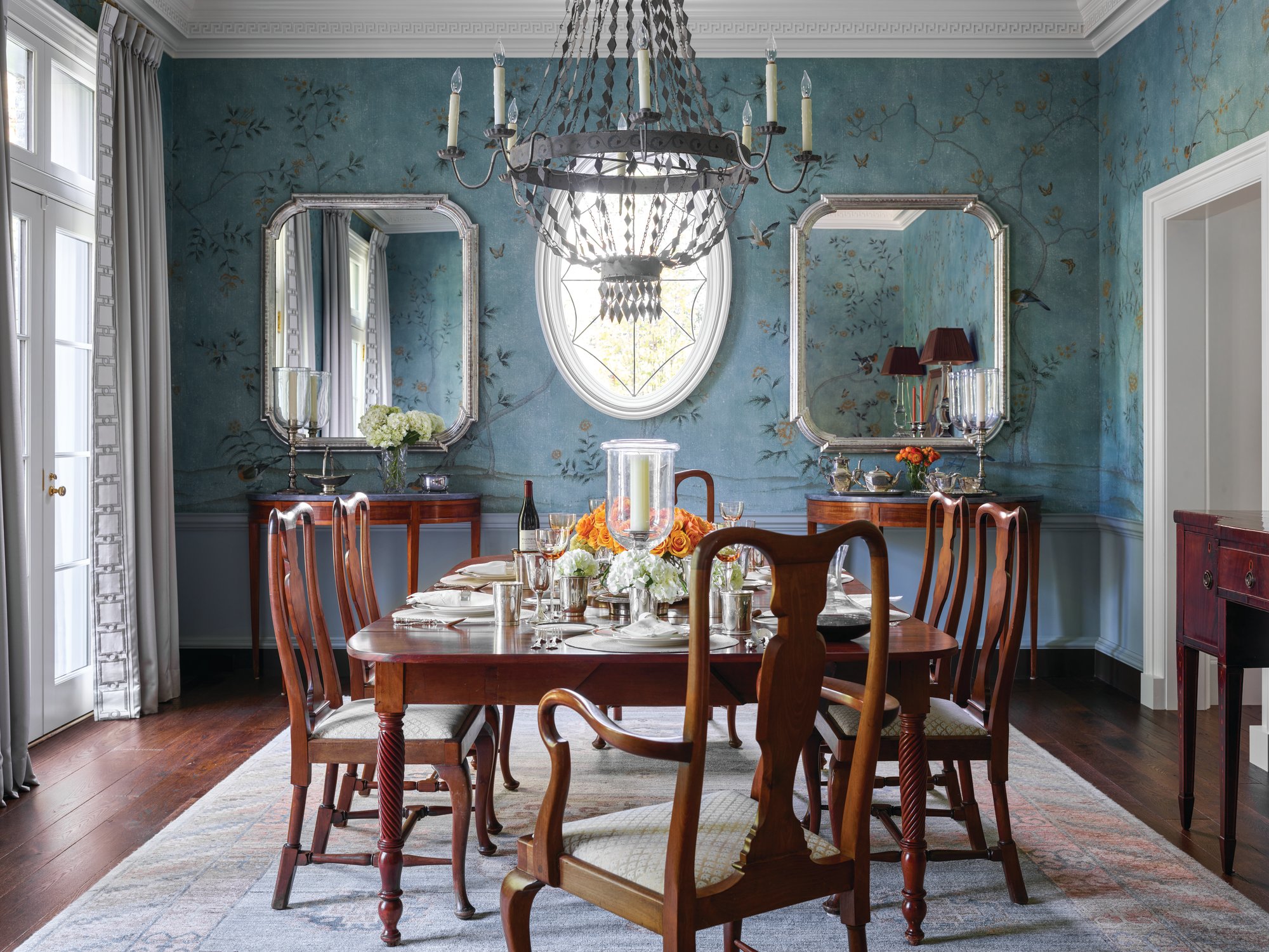 Dining room with blue hand-painted chinoiserie wallpaper, silver-framed mirrors flanking an oval window, a beaded iron chandelier, and a mahogany table set with candles and flowers