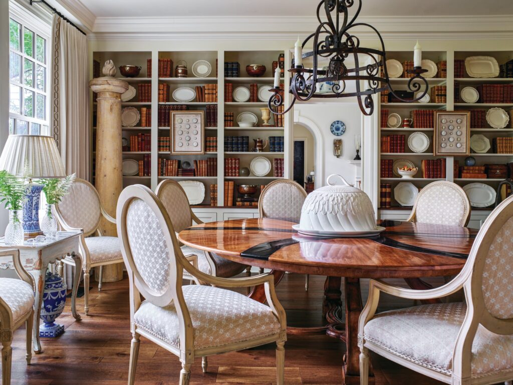 Library dining room lined with built-in bookshelves holding leather-bound books and creamware plates, with a round inlaid table, upholstered oval-back chairs, and an iron chandelier