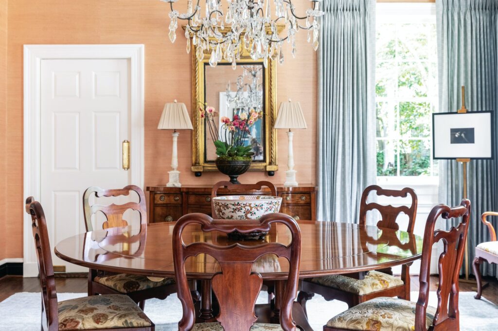 Dining room with blush grasscloth walls, a crystal chandelier over a round mahogany pedestal table, Queen Anne chairs, and a gilt mirror above a chest