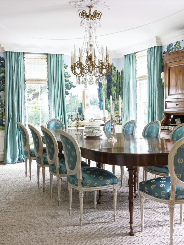 Dining room with scenic landscape mural wallpaper, aqua silk curtains, a gilt crystal chandelier, and a long mahogany table with blue upholstered Louis XVI chairs