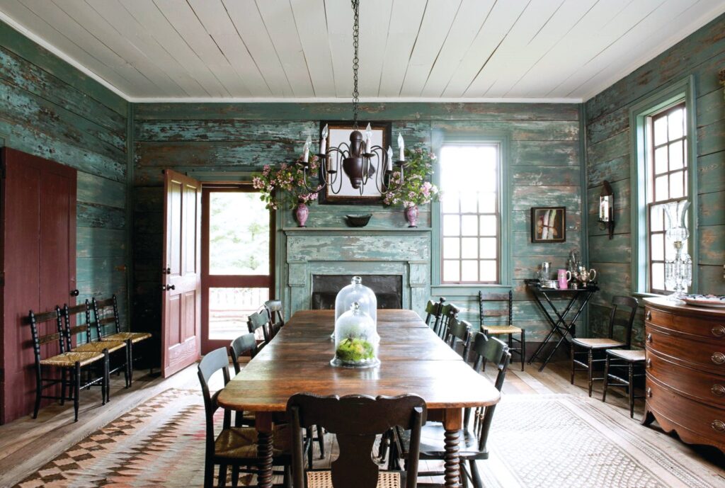 Rustic dining room with weathered green painted board walls, a long farmhouse table with rush-seat chairs, a mantel with flowers, and a bowfront chest
