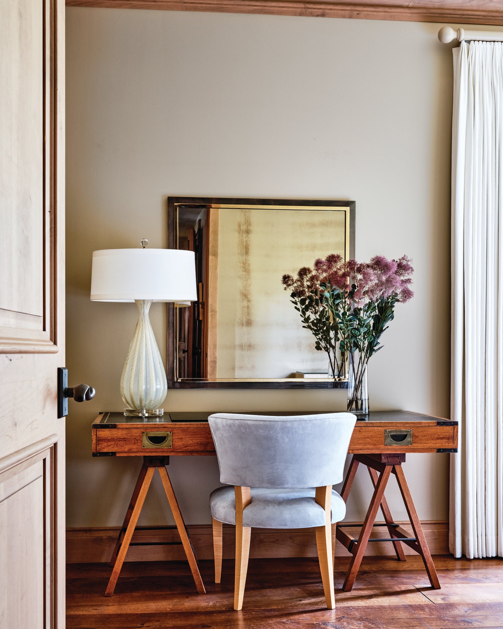 Campaign-style desk with brass-trimmed drawers on a sawhorse trestle base, paired with a gray upholstered chair, a glass lamp, and a mirror