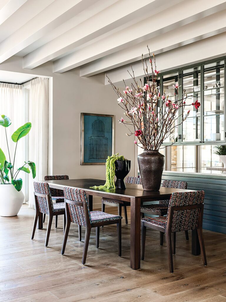 Dining room with a dark wood Huppé table, chairs in colorful pixel-pattern velvet, a tall vase of magnolia branches, and a beamed ceiling