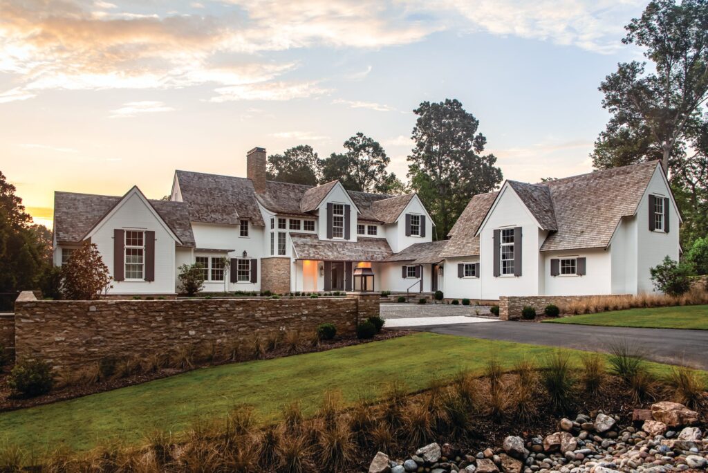 Front approach to a white European-style country house in Cary, North Carolina, with cedar shake roofs, dark shutters, and a stone wall, broken into a main house, garage, and guesthouse