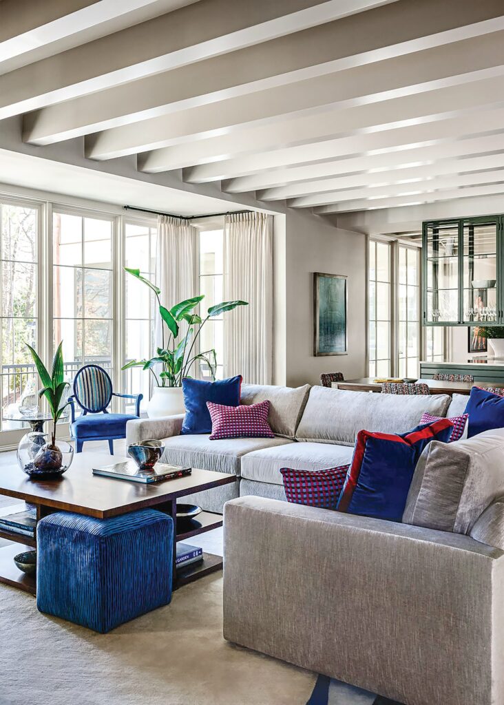 Living room with a gray sectional, blue velvet and red pillows, blue velvet ottoman cube, blue lacquered Baker chair, and tall windows with sheer drapes