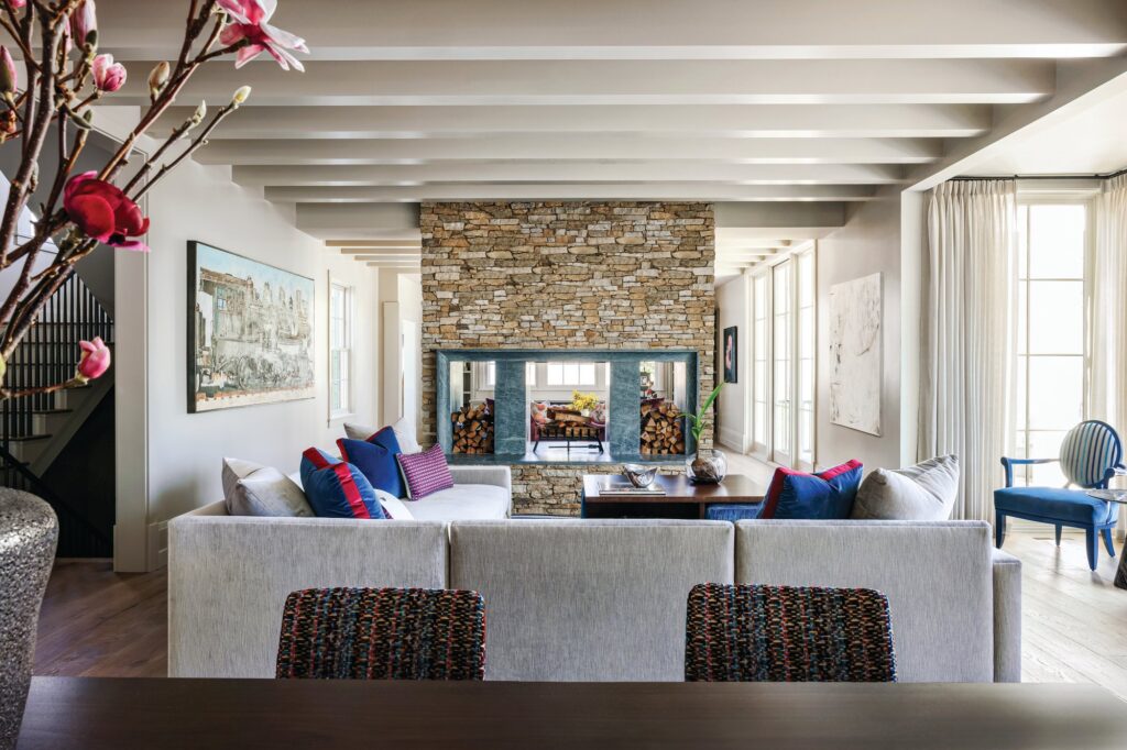 Open living room with a beamed white ceiling, a gray sectional with blue and red pillows, and a central double-sided stacked stone fireplace