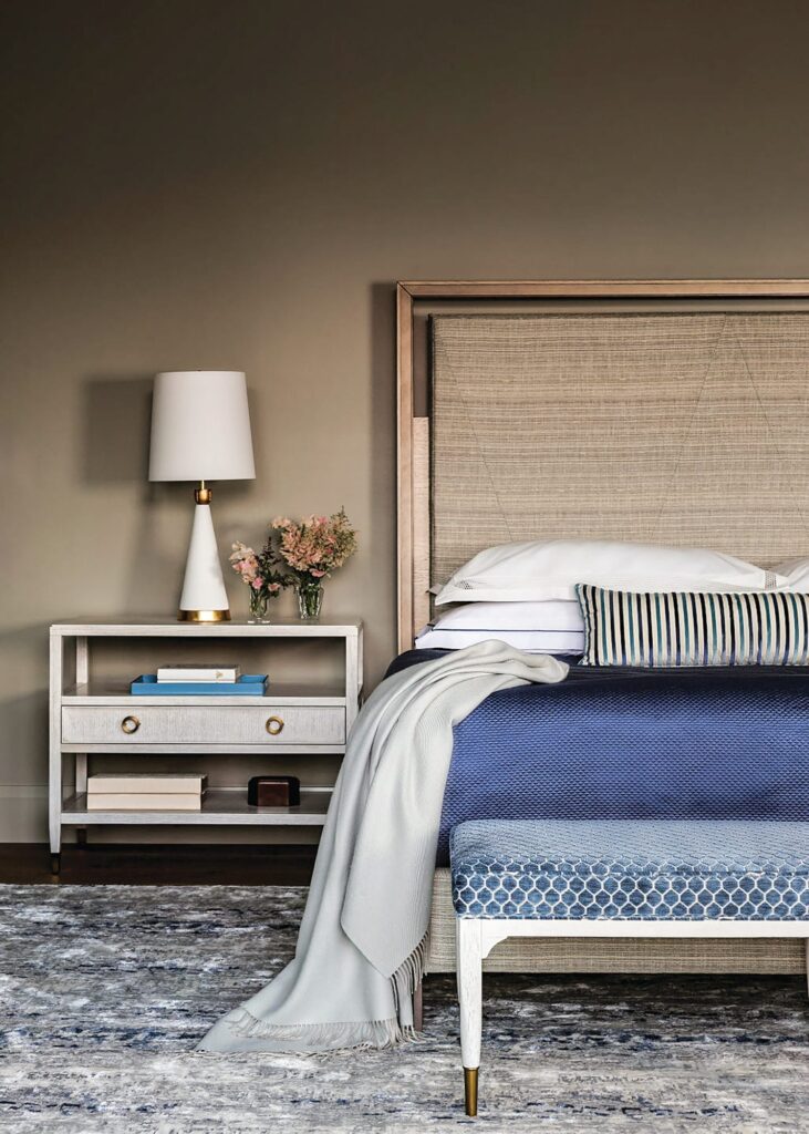 Primary bedroom with a Hurtado bed with woven headboard, blue bedding, a patterned bench, white nightstand, and taupe walls