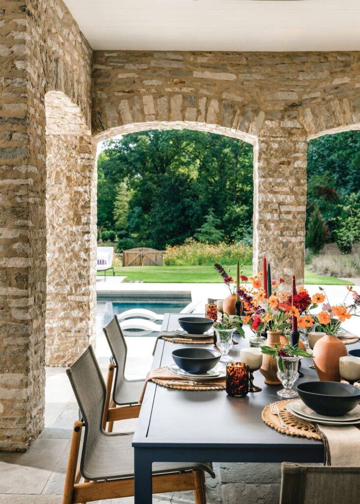 Covered outdoor dining under stone archways with a set table and orange flowers, overlooking the pool and gardens