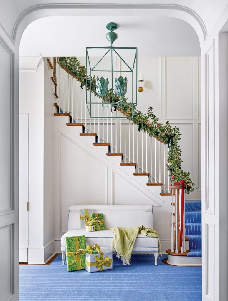 Grand foyer with a green-painted lantern chandelier, staircase garland with ribbons, and wrapped gifts on a blue runner