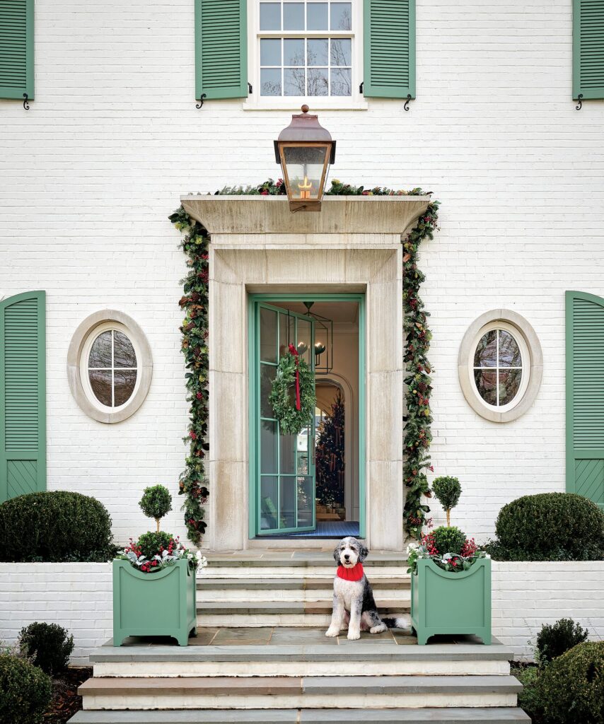 White brick home with green shutters and a green steel entry door dressed with garland, with a Bernedoodle in a red scarf on the steps