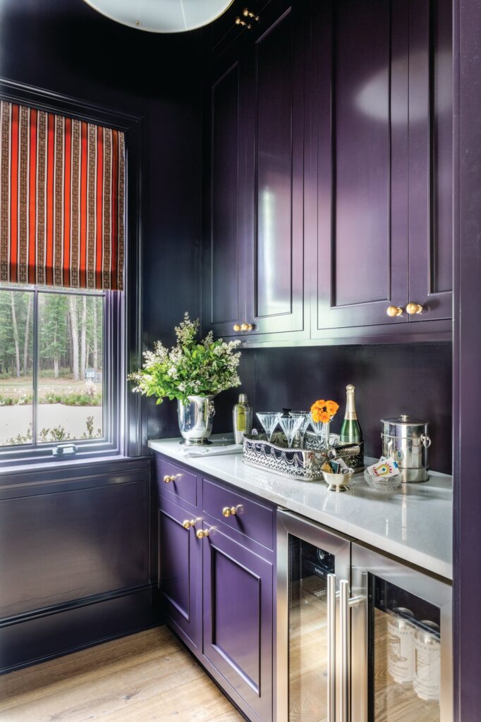 Butler’s pantry with aubergine lacquered cabinetry, a striped Roman shade, a beverage refrigerator, and a bar tray with glassware