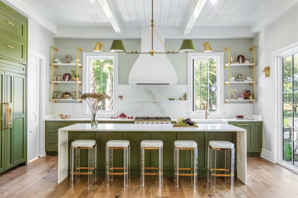 Kitchen with a green island painted in Farrow and Ball Bancha, five acrylic counter stools, a white plaster range hood, marble backsplash, and brass open shelving
