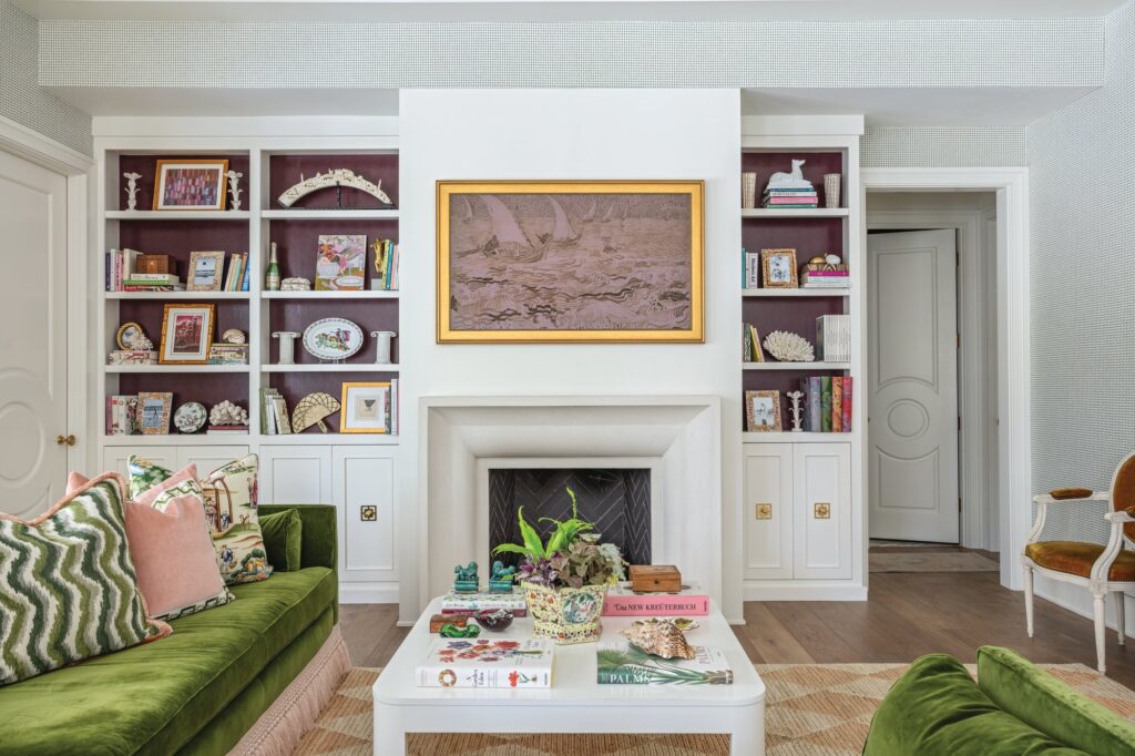 Living room fireplace flanked by built-in bookshelves with aubergine backs, a gilt-framed painting above the mantel, and green velvet sofas