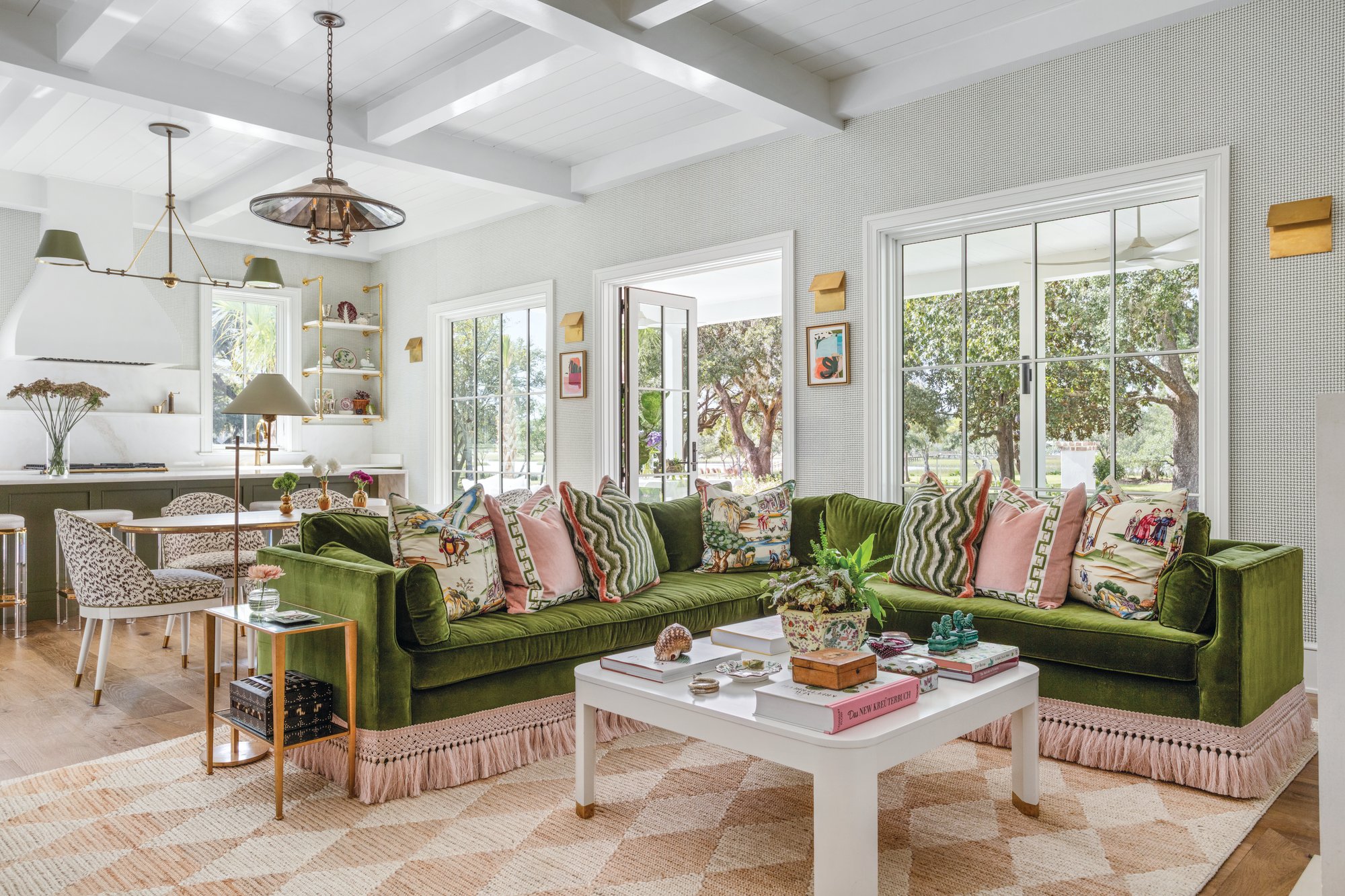 Living room with an olive green velvet sectional with a pink fringe skirt, patterned pillows, a white lacquered coffee table, and windows looking out to oak trees