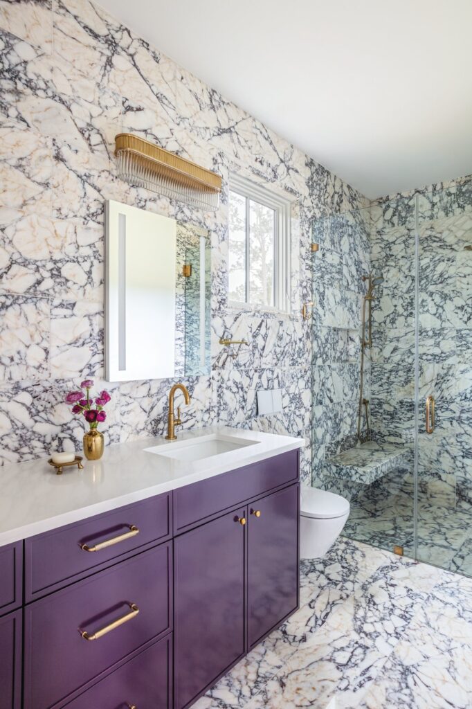 Primary bath clad floor to ceiling in Calacatta Viola marble with a purple vanity, brass faucet and sconce, and a glass shower