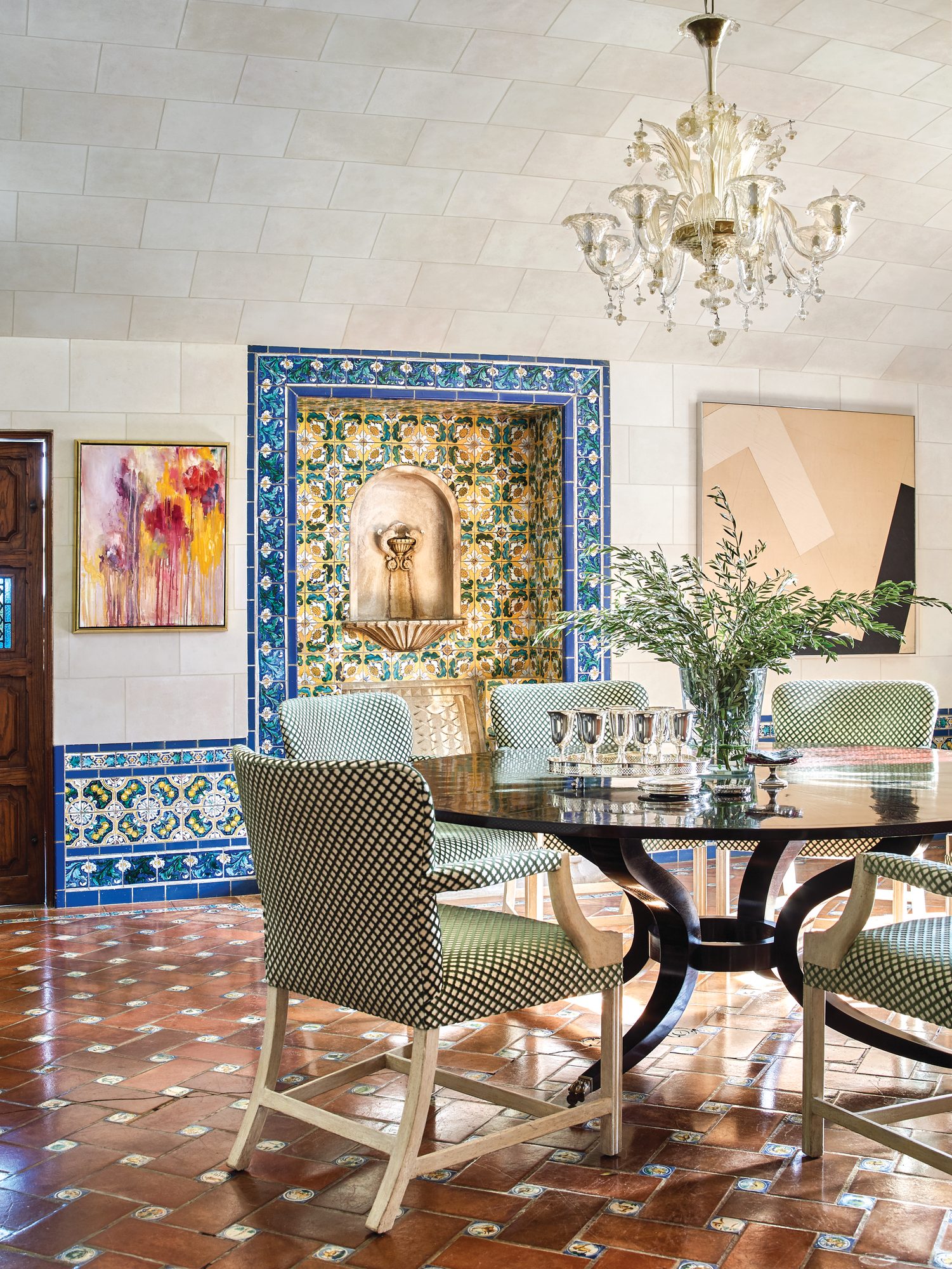 Dining room with a blue and gold tiled fountain alcove, stone-effect painted walls, Murano glass chandelier, round table, checked upholstered chairs, and contemporary paintings