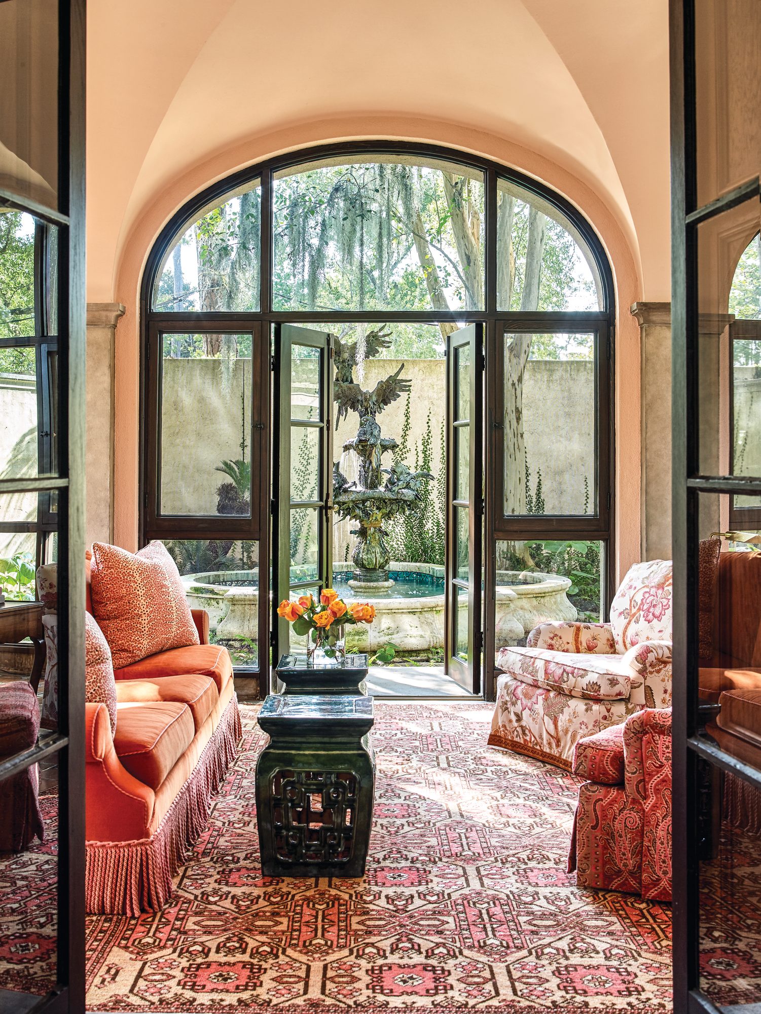 Sitting room with a coral velvet sofa, floral armchair, red patterned rug, and arched steel doors opening to a garden fountain
