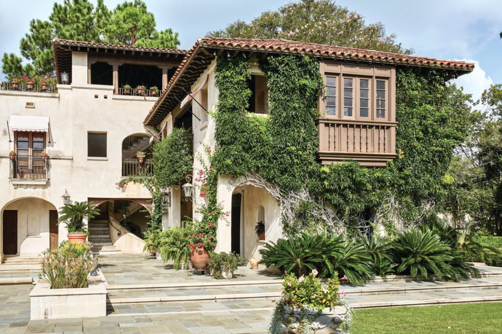 Ivy-covered stucco facade of a 1926 Spanish Revival home in Houston’s River Oaks with a red tile roof, carved wood balcony, arched openings, and a stone terrace with potted palms
