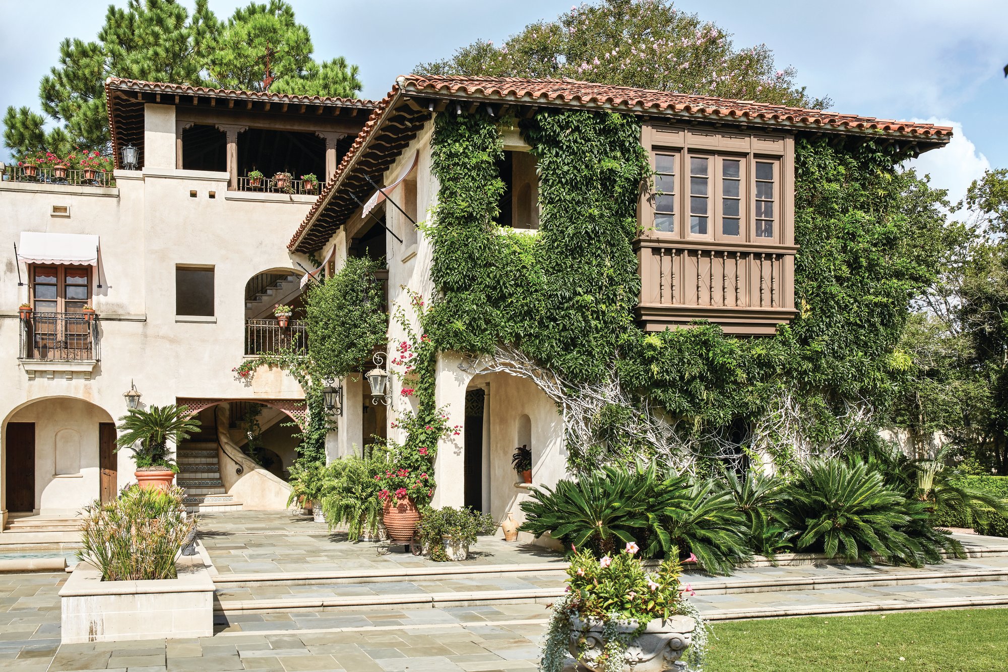 Ivy-covered stucco facade of a 1926 Spanish Revival home in Houston’s River Oaks with a red tile roof, carved wood balcony, arched openings, and a stone terrace with potted palms