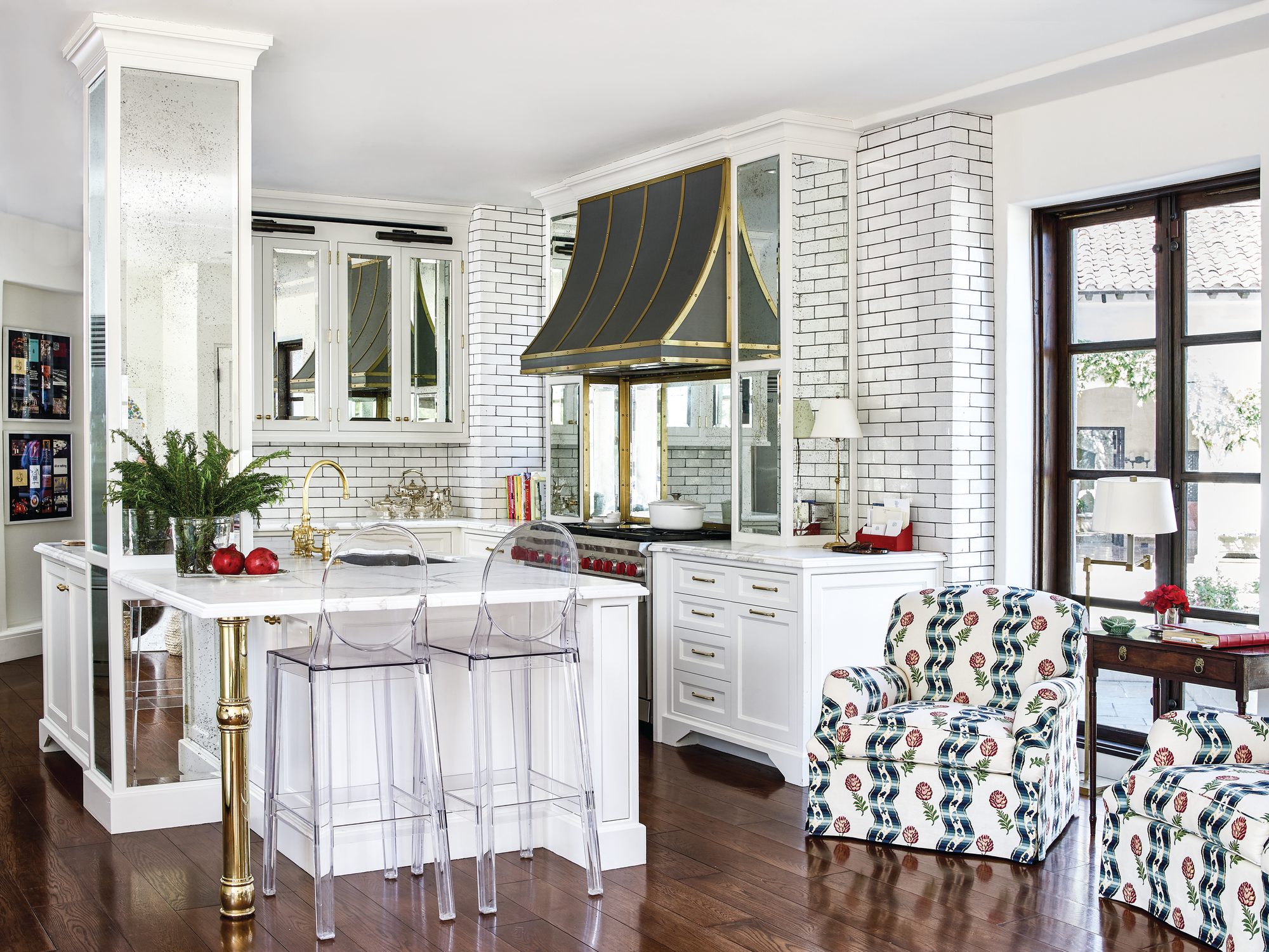 White kitchen with an antiqued mirrored column, brass island post, acrylic counter stools, dark brass-trimmed range hood, white tile walls with dark grout, and patterned armchairs