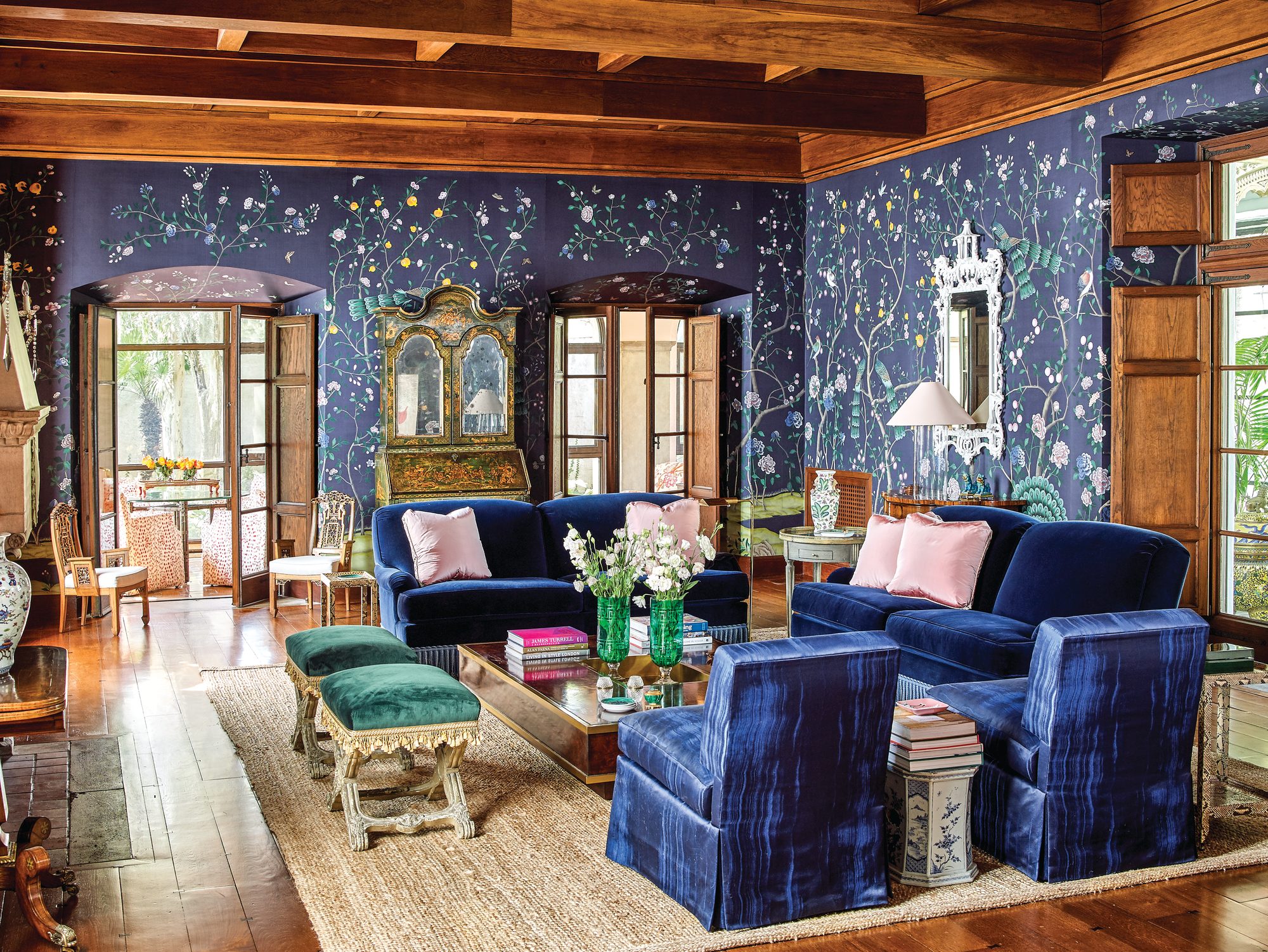 Living room with deep blue floral de Gournay wallpaper, beamed wood ceiling, blue velvet sofas with pink pillows, slipper chairs, and a natural fiber rug