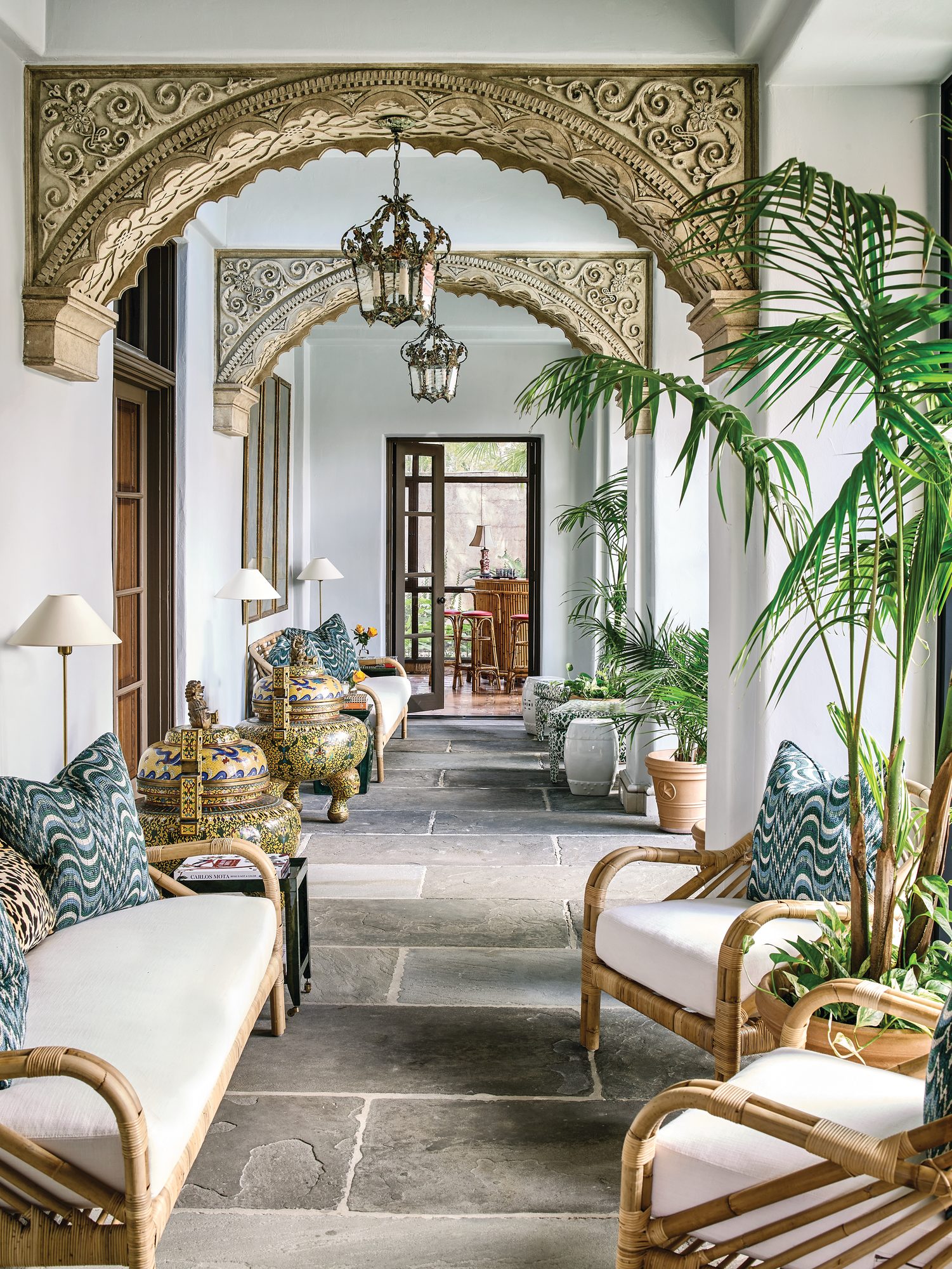 Loggia with carved stone arches, wrought iron lanterns, rattan seating with white cushions, flagstone floors, potted palms, and blue and gold ceramic urns